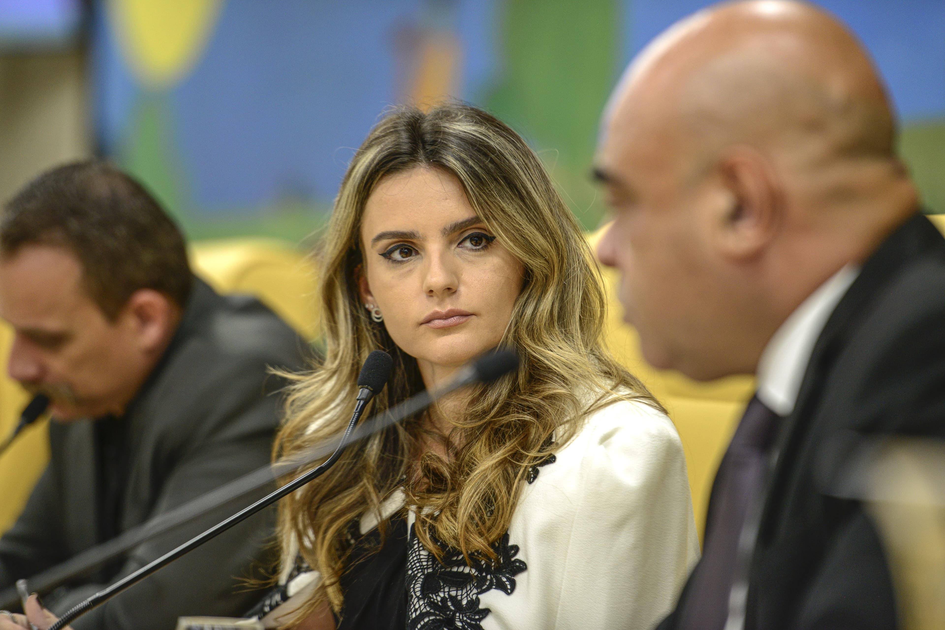 Amanda Vettorazzo, de blazer branco, ouve atenta entre dois homens numa mesa de reunião