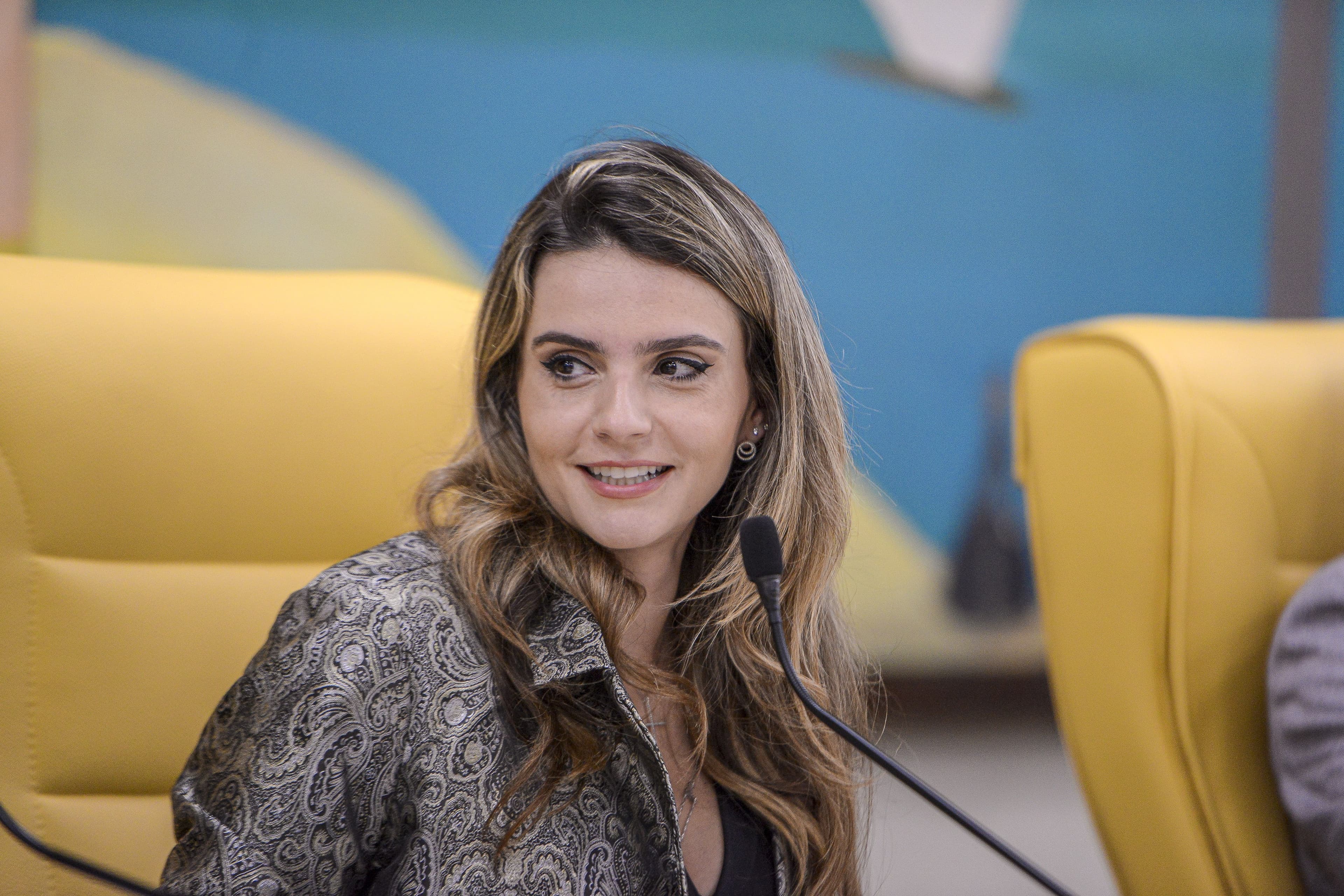 Amanda Vettorazzo sorri, sentada diante de um microfone, com um painel colorido ao fundo