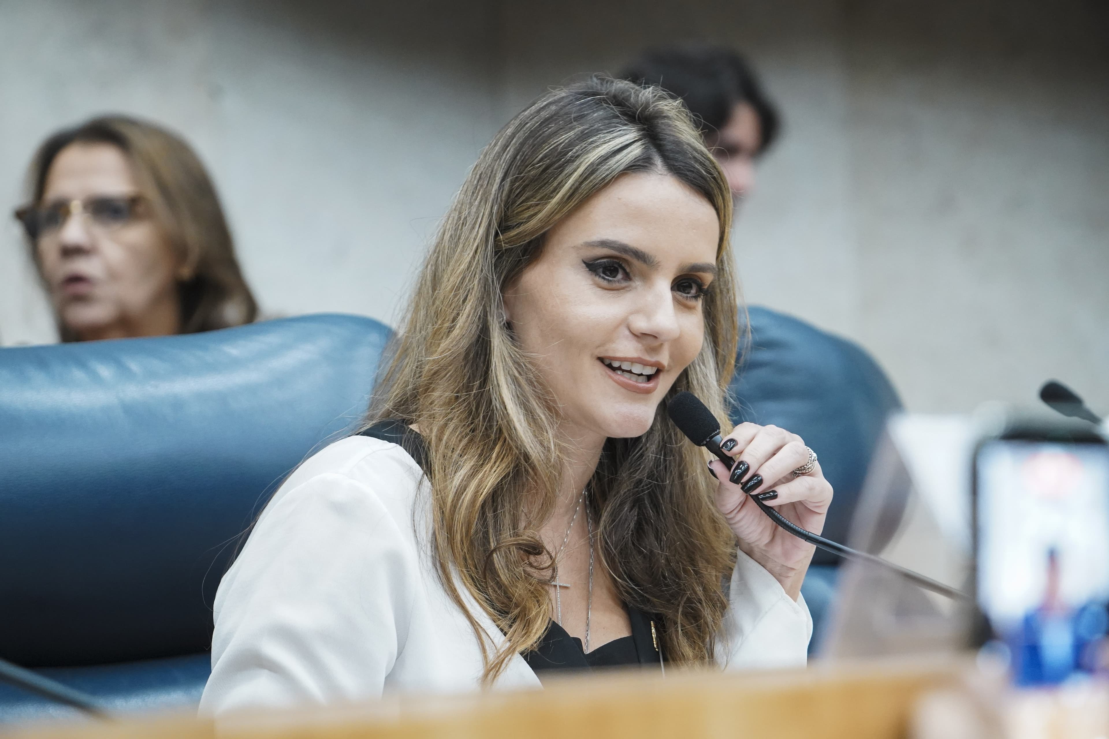 Amanda Vettorazzo, de blazer branco, sorri ao microfone numa sessão
