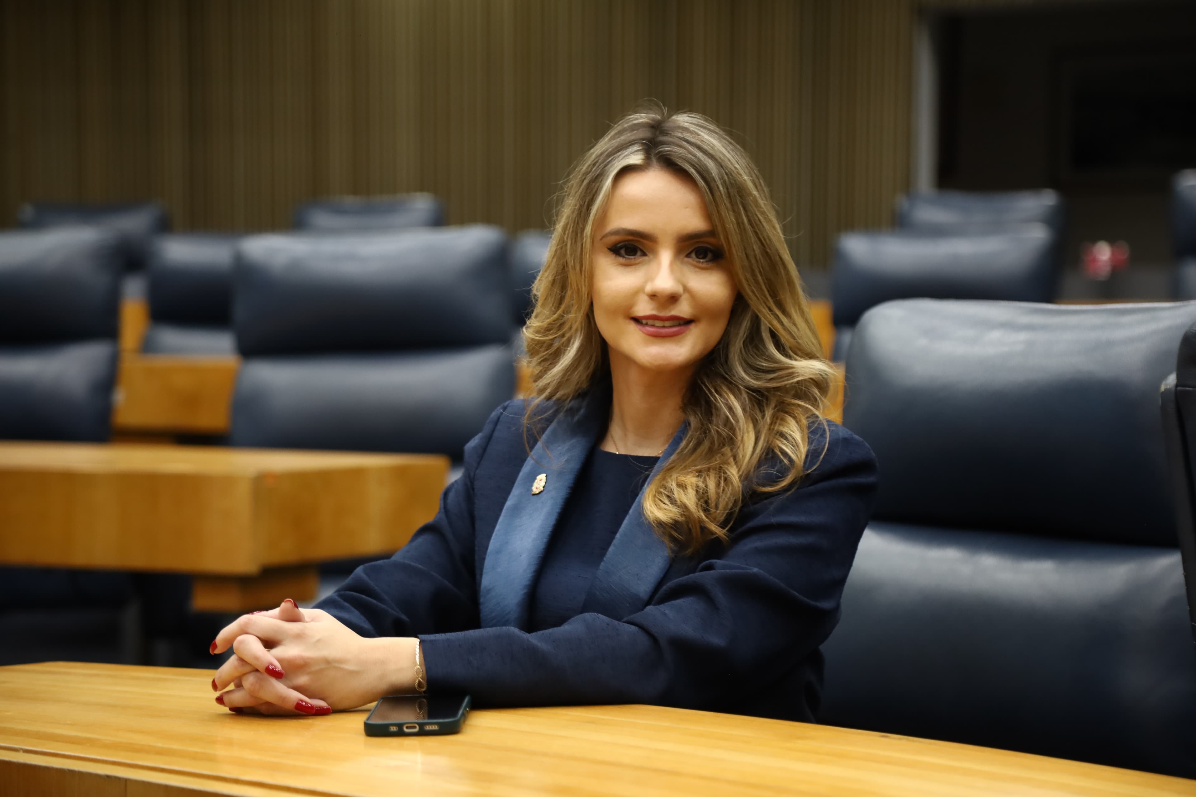 Amanda Vettorazzo, de blazer azul-marinho, sentada numa bancada do plenário da Câmara Municipal de São Paulo, olhando para a câmera